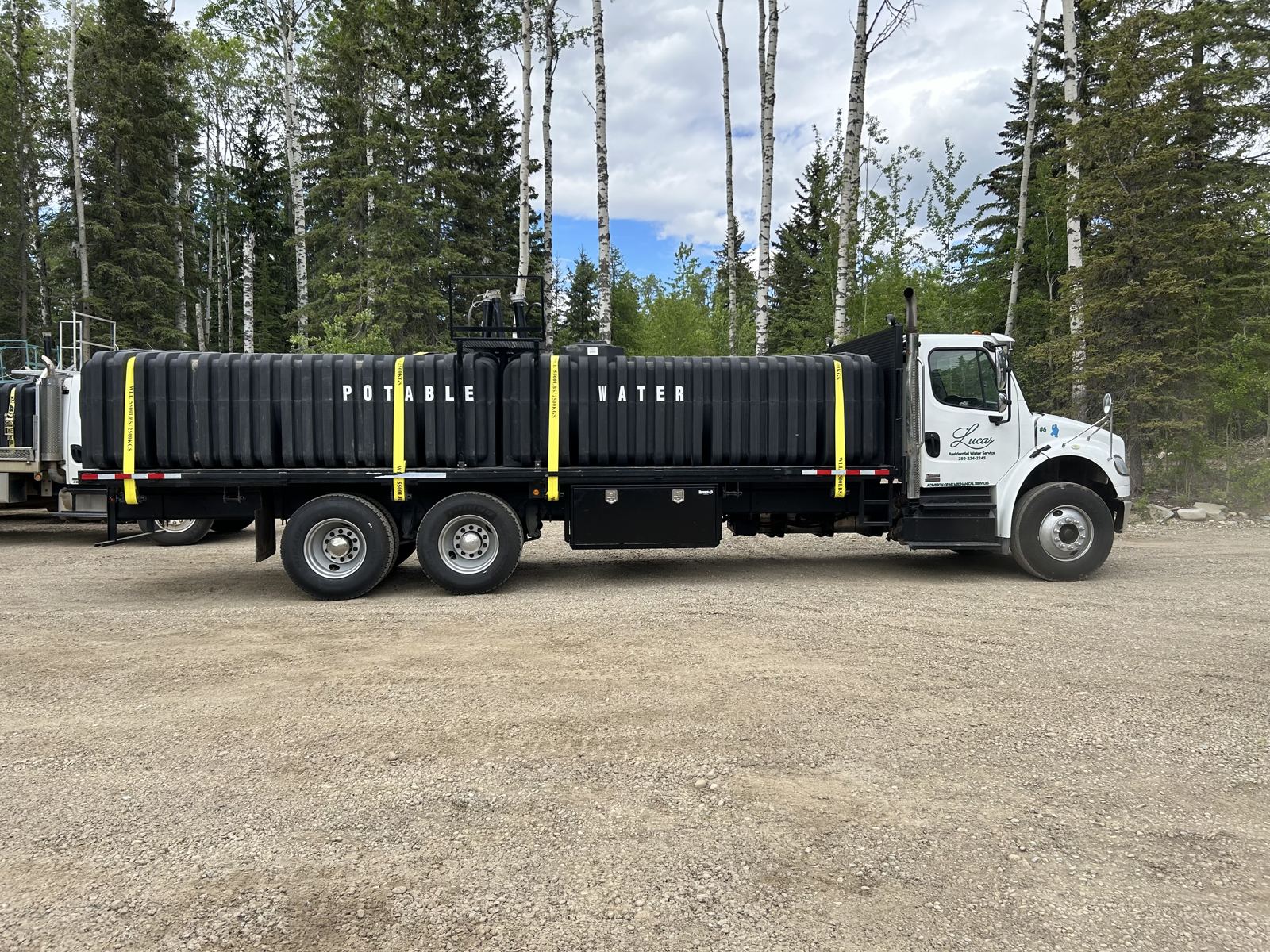 A white Lucas Residential Water Service tanker truck parked at the edge of a spruce forest